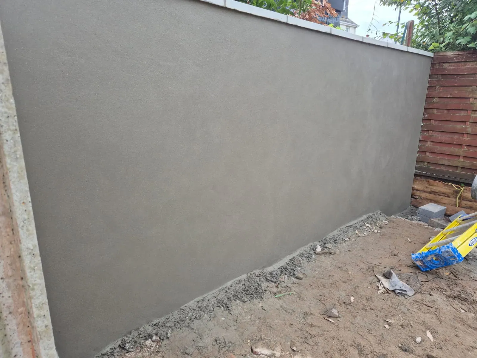 A freshly rendered grey outdoor wall, possibly a garden boundary, with new plasterwork visible and groundworks in progress.