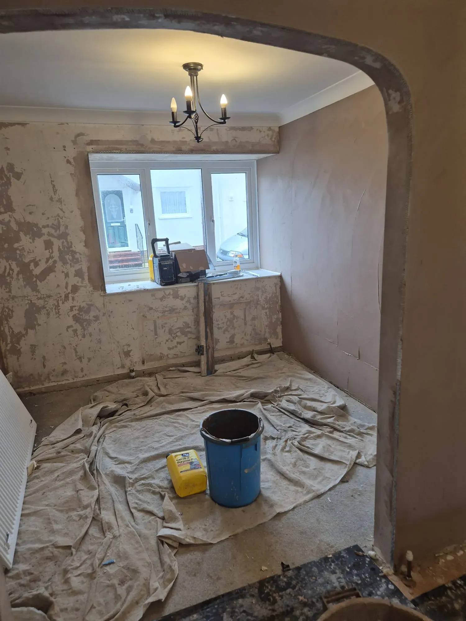 Interior room during renovation, featuring a newly plastered wall, stripped walls, an arched opening, and building materials on dust sheets. Represents home refurbishment and plastering services.